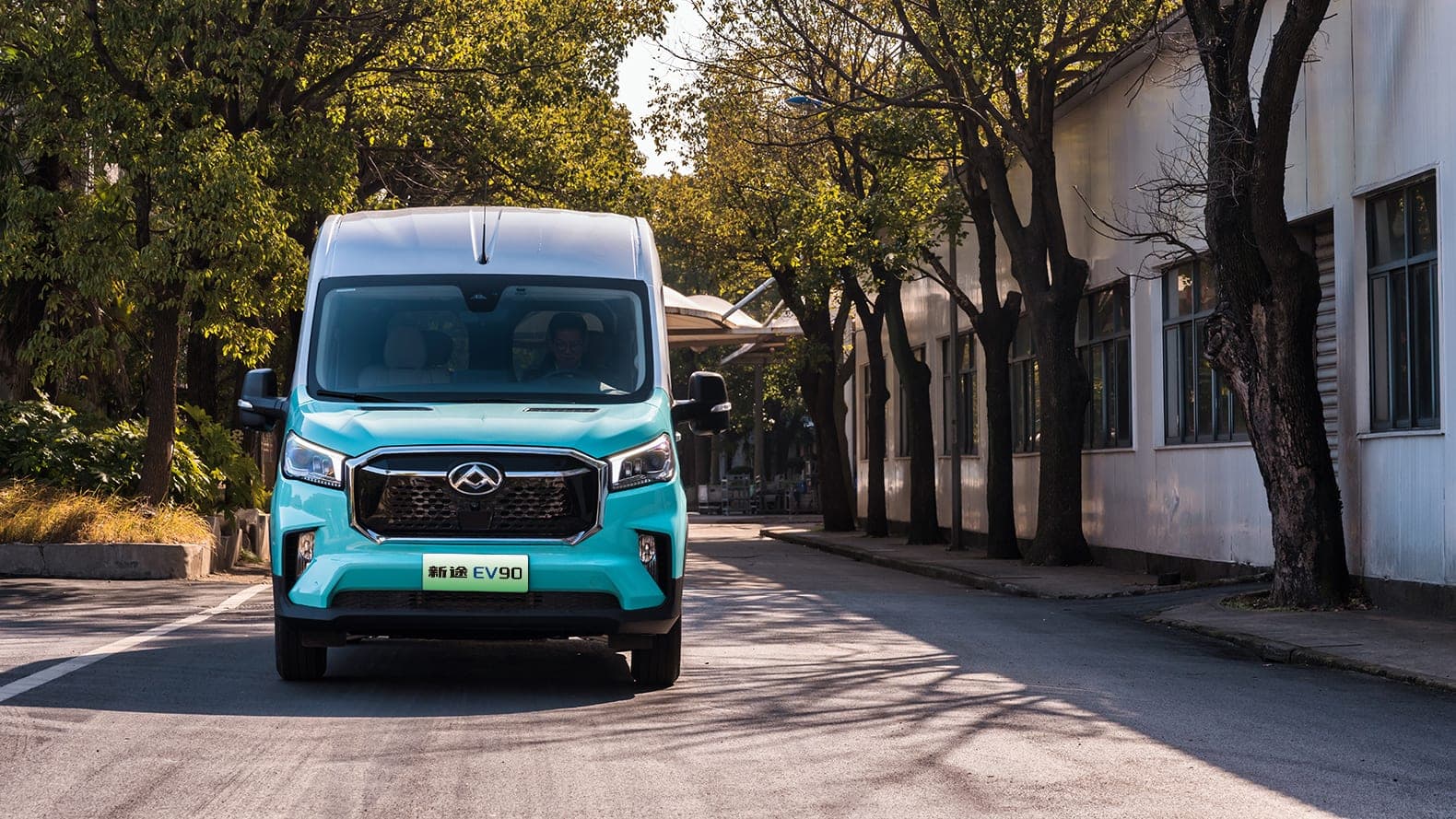 MAXUS EV90 2026 — فان كهربائي كبير وحافلة صغيرة من 9 إلى 15 مقعدًا للتصدير, منظر أمامي في شارع تحفه الأشجار